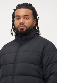 Mand med skæg og dreadlocks iført en sort quiltet jakke med høj krave, der kigger direkte ind i kameraet mod en ensfarvet baggrund.
