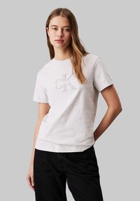 Mujer joven con cabello largo que lleva una camiseta gris claro de Calvin Klein y pantalones negros, de pie frente a un fondo gris liso.
