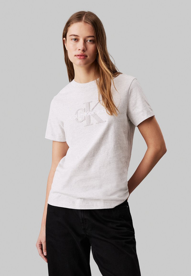 Mujer joven con cabello largo que lleva una camiseta gris claro de Calvin Klein y pantalones negros, de pie frente a un fondo gris liso.