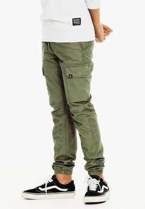 Pantalons cargo verts avec des poignets élastiques, plusieurs poches et un tissu texturé. Portés avec des baskets noires avec des accents blancs.