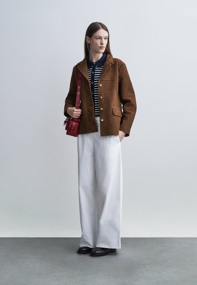 Γυναίκα που φορά καφέ σακάκι με κουμπιά, μπλε πουκάμισο με ρίγες, λευκό παντελόνι φαρδιάς γραμμής, μαύρα παπούτσια, και κρατά μικρό κόκκινο τσαντάκι στον ώμο.