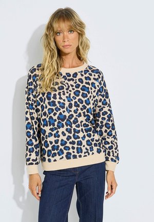 Mujer con pelo largo y rubio que lleva un suéter beige con estampado de leopardo azul y negro y vaqueros azul oscuro sobre un fondo liso.