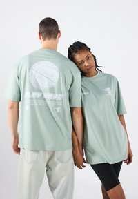 Piparmētru zaļas oversized t-krekli ir ar grafiskām printēm, tostarp basketbola attēlu un tekstu "SPĒLĒ CIEŠI", apvienoti ar gaišajiem džinsiem un melnajām šortiem.