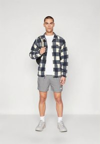 Ζακέτα από φλις με γιακά και μοτίβο σε ναυτικό μπλε και κρεμ καρό, λευκό t-shirt, γκρι σορτς και ανοιχτόχρωμα αθλητικά παπούτσια, κρατώντας ένα σακίδιο.