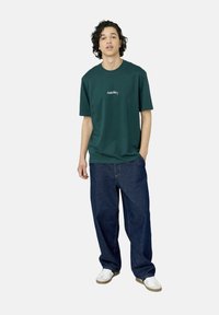 T-shirt en coton vert foncé avec un col rond et du texte brodé blanc, associée à un jean bleu en denim ample et des baskets blanches.