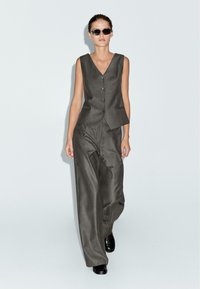 Gilet grigio a righe sottili e pantaloni a gamba larga, con scollatura a V profonda e chiusura a bottoni. Abbinato a scarpe nere e occhiali da sole.