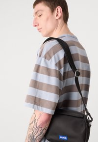 Sort nylon crossbody taske med lynlås, logo-patch og justerbar rem, båret over en grå og brun stribet T-shirt.