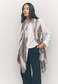 Sciarpa leggera con design strutturato e motivi in sfumature di beige e marrone, con bordi sfrangiati, avvolta intorno a una camicia bianca.