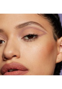 Gros plan sur un visage avec un teint chaud, featuring un fard à paupières violet doux, un design d'eyeliner prononcé, et des lèvres nues mates avec une texture subtile.