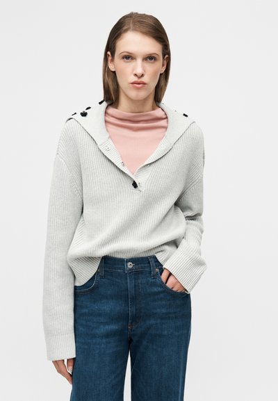 Ανοιχτό γκρι πλεκτό πουλόβερ με κουκούλα, με διακοσμητικά κουμπιά και βαθύ V-λαιμόκοψη. Φορεμένο πάνω από ένα ροζ τοπ και συνδυασμένο με σκούρο μπλε τζιν.
