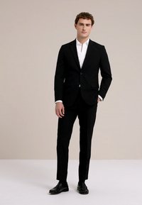 Jonge man staand, gekleed in een zwart pak met een wit overhemd en zwarte nette schoenen, tegen een effen beige achtergrond.