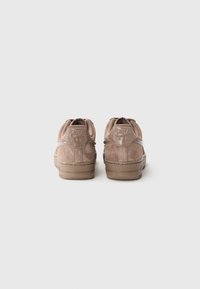 Baskets en suède beige avec des talons arrondis, semelles en caoutchouc et un accent de logo brillant au niveau du talon. Présente un laçage minimal et des détails texturés.