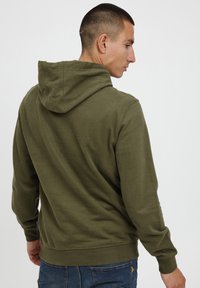 Olive green cotton hoodie with a drawstring hood, ribbed cuffs and hem, minimal stitching, and a relaxed fit. Worn with blue jeans.