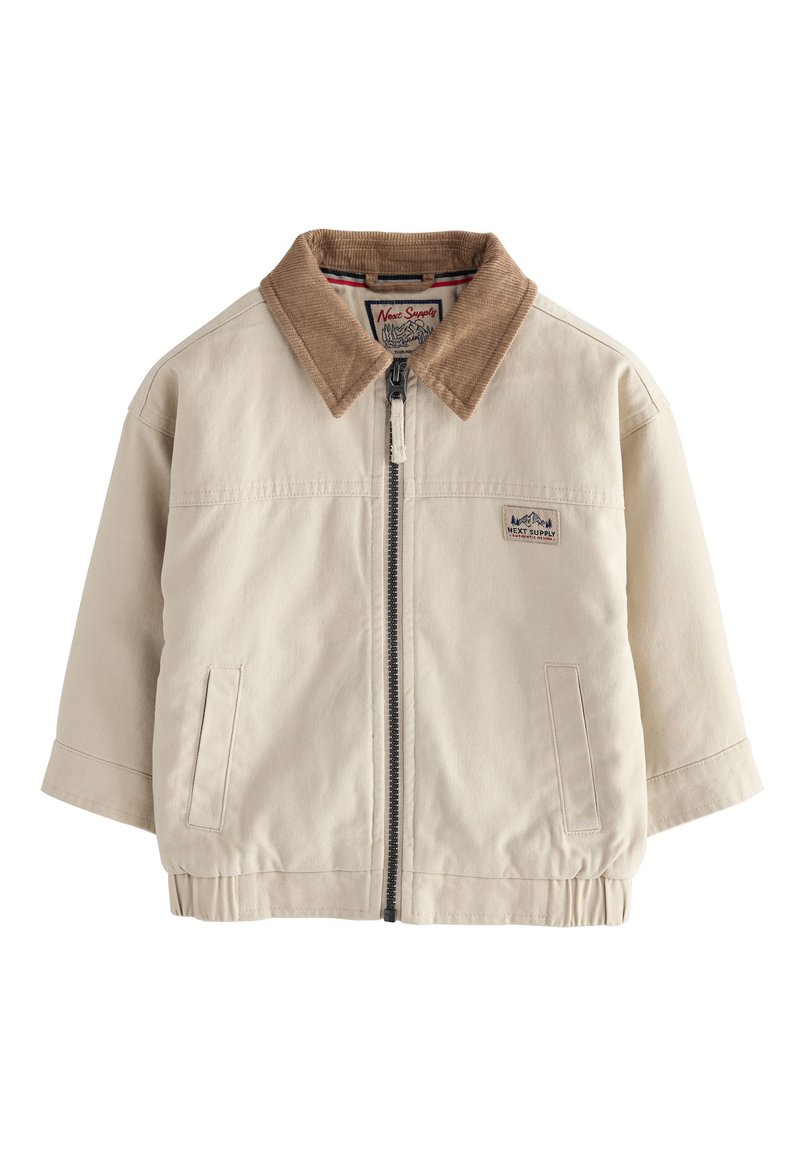 Bézs dzseki barna kordbársony gallérral, elülső cipzárral és rugalmas szegéllyel. Oldalzsebekkel és a mellkason egy kis logóval rendelkezik.