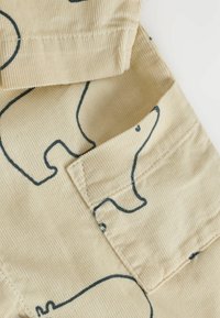 Beige kordfløyel med svart omriss av isbjørntegninger och en detaljerad närbild av en framfickas lap på ett plagg.