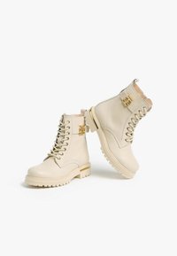 Bottes cheville en cuir beige avec bout arrondi, fermeture à lacets, col rembourré, accent de ferrure dorée et texture de semelle extérieure robuste.