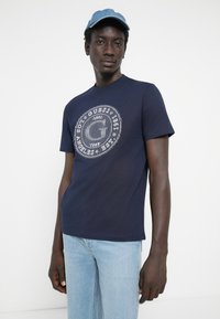 Mørkeblå bomulds t-shirt med et cirkulært hvidt grafisk design på fronten. Modellen har lyseblå jeans og en denimkasket på.