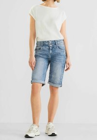 Light blue denim shorts with rolled cuffs, featuring a button and zipper fly closure, five pockets, and slightly distressed areas. Worn with a white top.