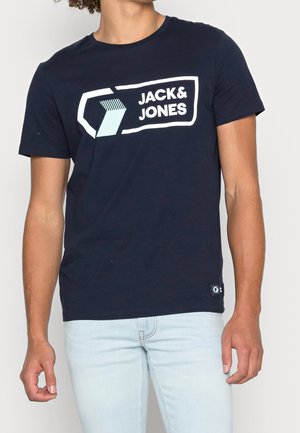 Mężczyzna ubrany w granatowy t-shirt z logo Jack & Jones i jasnoniebieskie dżinsy, stojący na tle jednolitego białego tła.