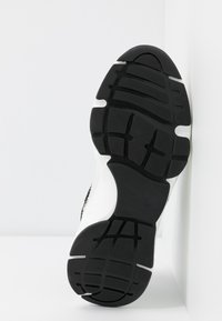 Semelle de chaussure de sport noire et blanche avec des crampons texturés, présentée sur un fond blanc et appuyée contre une surface blanche.