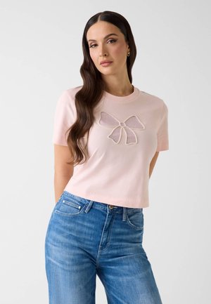 Vrouw met lang, donker golvend haar, gekleed in een lichtroze top met korte mouwen en decoratieve strik, gecombineerd met een blauwe high-waisted jeans, staand tegen een effen achtergrond.