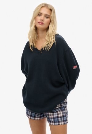 Pull en maille navy avec un col en V, coupe oversize et base côtelée ; associé à un short à carreaux aux couleurs bleues, rouges et crème.