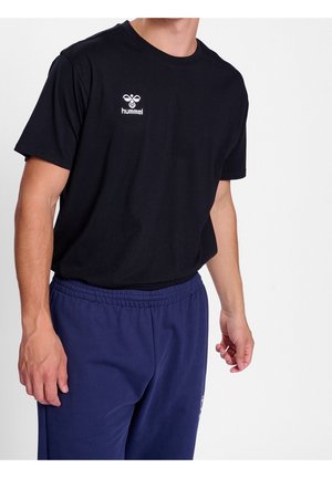 Mann trägt schwarzes Hummel-T-Shirt und dunkelblaue Jogginghose, steht vor schlichtem weißem Hintergrund.