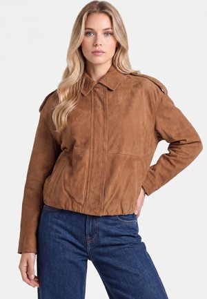 Femme blonde aux ondulations lâches portant une veste en daim marron et un jean bleu, posant avec une main sur la hanche sur fond blanc.