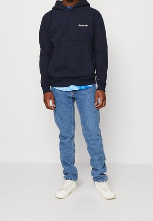 Mężczyzna w granatowej bluzie z kapturem z małym białym napisem, pod spodem niebieska koszulka tie-dye, jasnoniebieskie dżinsy i białe trampki, stojący na tle szarej ściany.