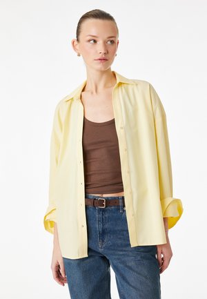 Młoda kobieta w luźnej, żółtej koszuli zapinanej na guziki, nałożonej na brązowy top i niebieskie dżinsy, patrząca w bok na tle białego tła.