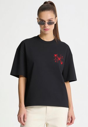 Frau mit braunen Haaren im Pferdeschwanz, die kleine rechteckige Sonnenbrillen trägt, ein schwarzes übergroßes T-Shirt mit rotem Tic-Tac-Toe-Herz-Design und hellen Hosen.