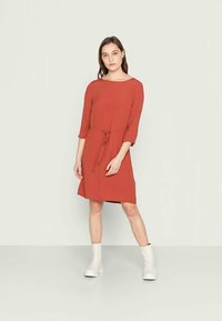 Vestido vermelho com um corte solto, mangas três quartos e uma cintura atada. Tecido liso, até ao joelho, combinado com botas brancas de tornozelo.