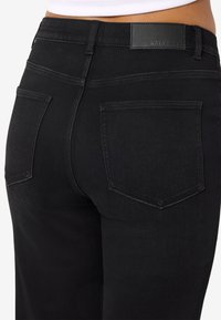 Približnji posnetek osebe, ki nosi črne kavbojke z visokim pasom, zadnjimi žepi in usnjenim znamčnim znakom z napisom »NOISY MAY«.