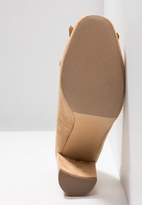 Chaussure à talon haute en suède beige avec une semelle en caoutchouc texturée, montrant un bout rond et un design sans couture sur le côté, positionnée à la verticale.