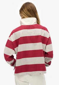 Maglia a maniche lunghe a righe rosse e grigie con collo alto, vestibilità rilassata e dettagli di cuciture visibili su un tessuto morbido.