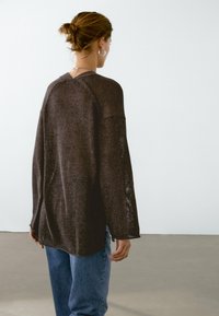 Woman with brown hair in a bun wearing silver hoop earrings, a loose dark brown sweater, and blue jeans against a plain light background.