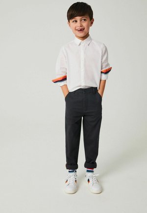 Camicia bianca con maniche arrotolate, caratterizzata da strisce rosse, arancioni e blu, abbinata a pantaloni neri e sneakers bianche.