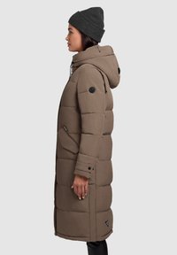 Dlhá hnedá páperová bunda s kapucňou, bočnými vreckami a okrúhlym logom na rukáve. Hladká textúra a prešívaný vzor.