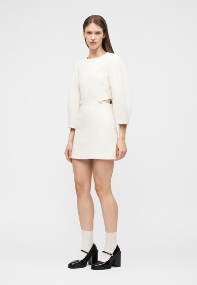 Γυναίκα που φοράει λευκό μίνι φόρεμα με υφή, μακρυά μανίκια και κοψίματα στο πλάι, συνδυασμένο με λευκές κάλτσες και μαύρα παπούτσια Mary Jane με χοντρό τακούνι.