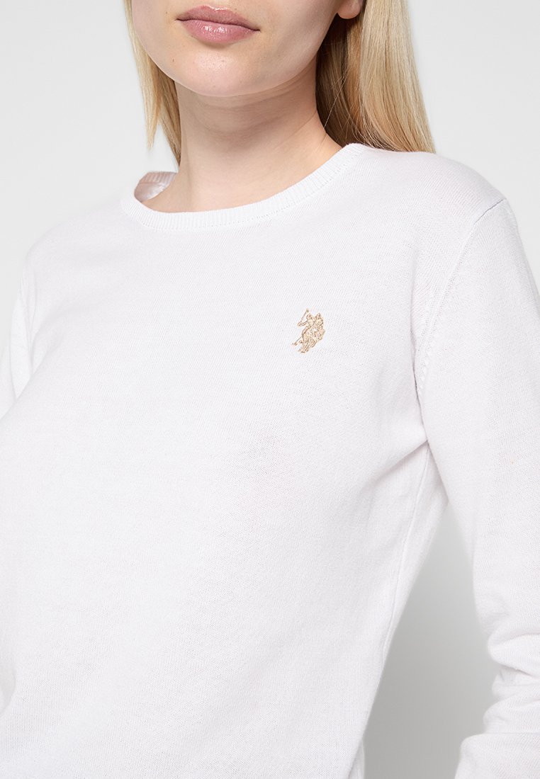 Frau in einem weißen Langarmoberteil mit einem kleinen, goldenen, gestickten Logo eines Pferdes und Reiters auf der linken Brust.