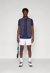 Marineblaues ärmelloses Hemd über einem Poloshirt mit roten Akzenten, weiße Shorts und Sportschuhe; hält einen Tennisschläger.