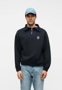 Άνδρας με ανοιχτό μπλε καπέλο, μαύρο φούτερ με φερμουάρ στο λαιμό και λογότυπο, και μπλε τζιν, στέκεται μπροστά σε λευκό φόντο.