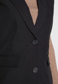 Black vest with a tailored design, featuring a notched lapel, double buttons, and a smooth texture. Layered over a ribbed brown top.