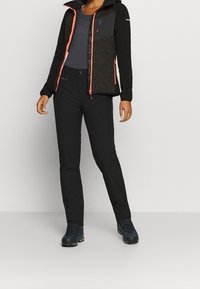 Schwarze Softshell-Jacke mit orangefarbenen Akzenten und einer strukturierten Platte, kombiniert mit schwarzer Hose und dunklem Schuhwerk. Das Outfit ist funktional und praktisch.