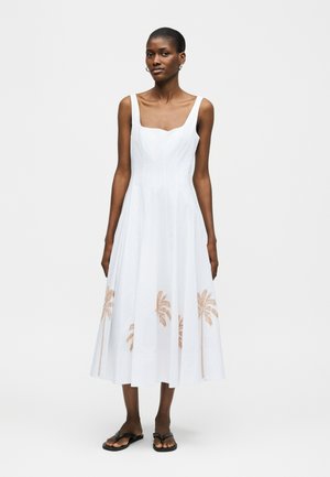 Γυναίκα που φοράει λευκό μίντι φόρεμα χωρίς μανίκια με τετράγωνο ντεκολτέ και χρυσά σχέδια με φοίνικες κατά μήκος της φούστας, συνδυασμένο με μαύρα σανδάλια.