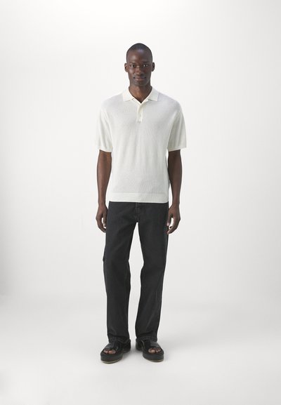 Polo shirt crème à manches courtes avec un motif tricoté texturé, associé à un jean en denim foncé et des sandales noires.