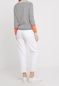 Top a maniche lunghe a righe con righe bianche e nere, polsini arancioni; indossato con jeans bianchi a vita alta e sneakers bianche, che mostrano un look casual.