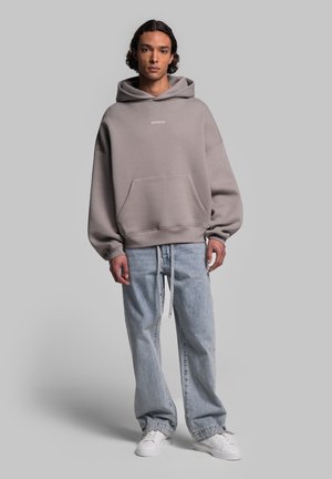 Oversized grey hoodie with a front pocket and white logo, paired with light blue baggy jeans and white sneakers. Minimalist design.