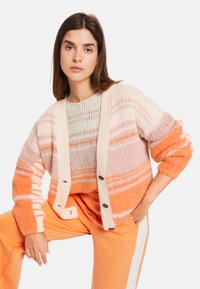 Gestreifter Cardigan in Pink- und Orange-Tönen mit kremfarbenem Knopfverschluss, weiten Ärmeln und strukturiertem Strickdesign, über einem leichten Pullover getragen.