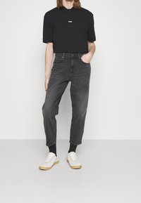 Polo nero con logo, abbinato a jeans grigi scuri a gamba stretta e sneakers bianche. L'outfit presenta un design semplice e pulito.